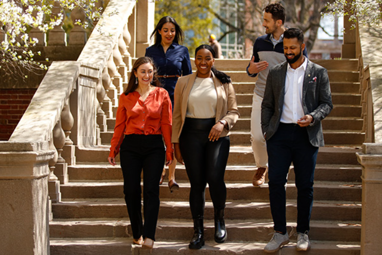 Five students-two men and three women--walk down steps on campus laughing and talking on a sunny day